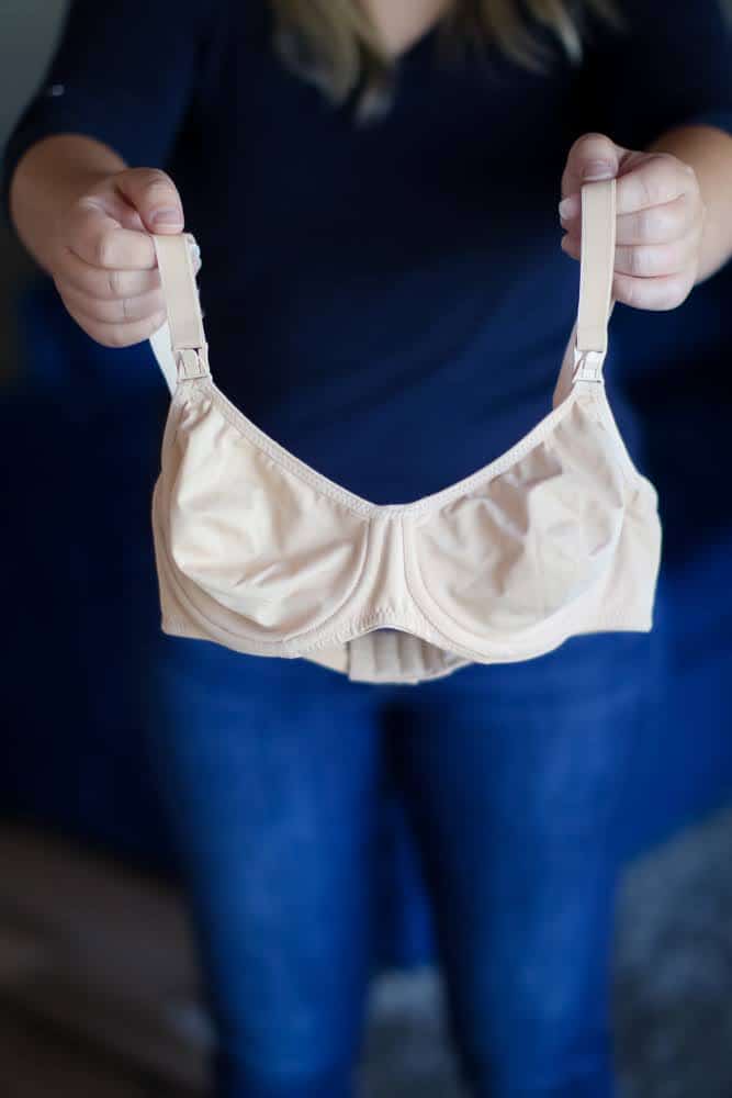 A woman in a blue t-shirt and denim jeans holds a nude nursing underwired bra with front clips