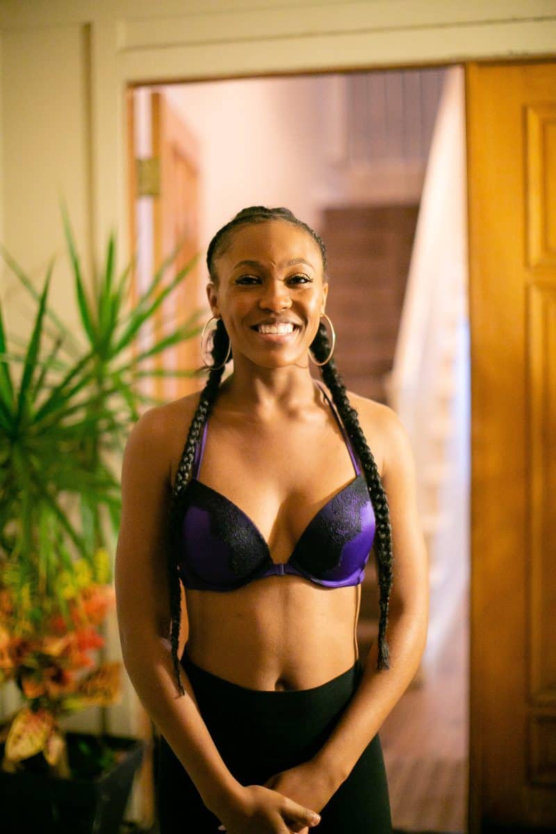 A woman in a purple plunge bra with black lace wearing black leggings stands near the brown door