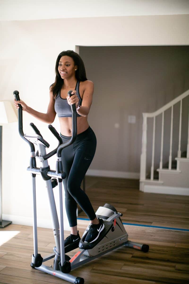Woman in a black sports bra and black leggings wearing black rubber shoes using an elliptical machine inside the house