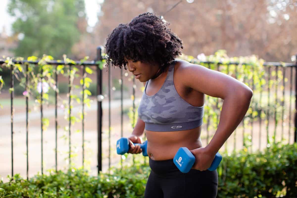 Woman in gray sports bra with a camouflage design and black leggings using blue dumbells outside the house