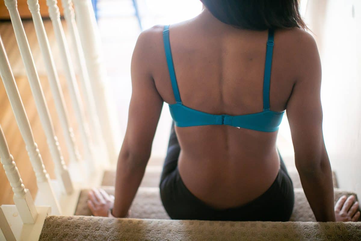 A woman shows the bra hooks of a blue bra with adjustable straps wearing black leggings and seated on the stairs