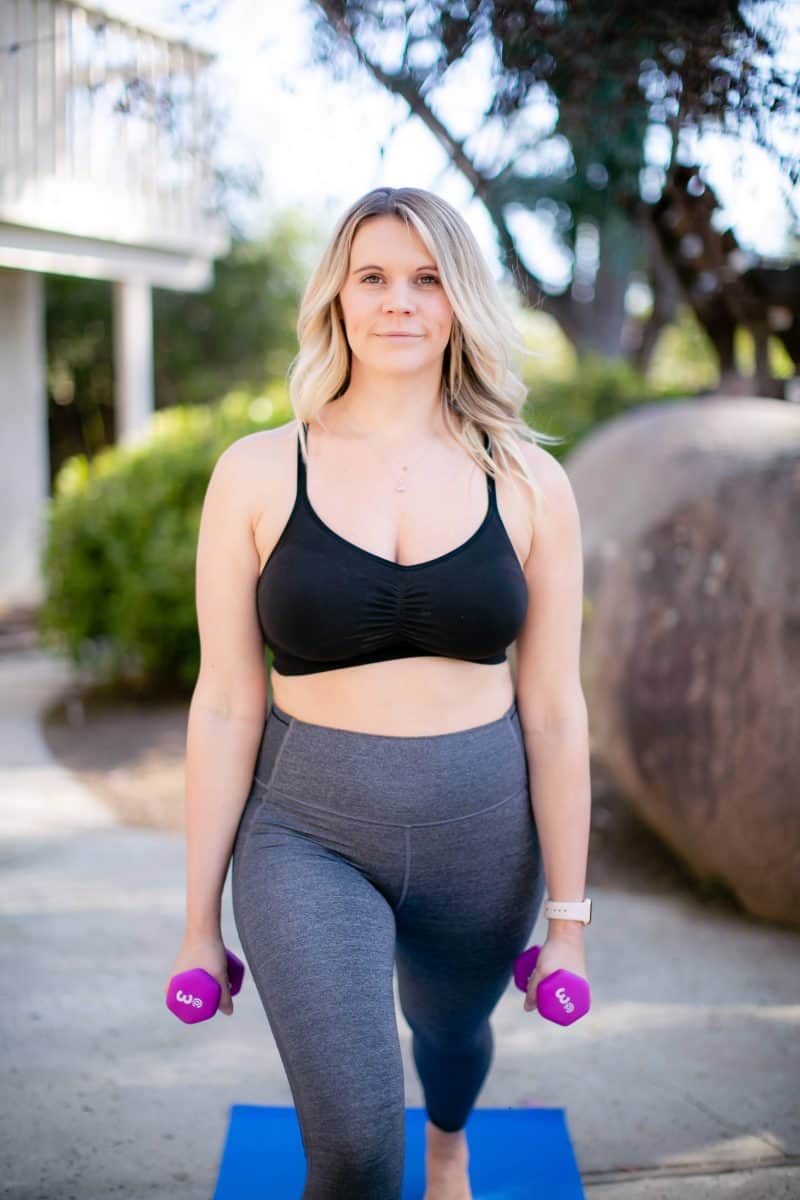 Woman in a black sports bra and gray leggings exercising using a dumbbell outside the house