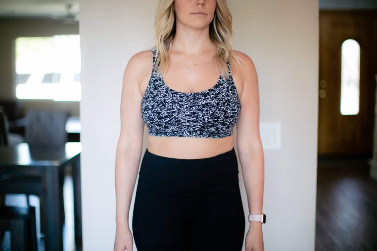 A black and white sports bra and black leggings are worn by a woman standing near a white wall