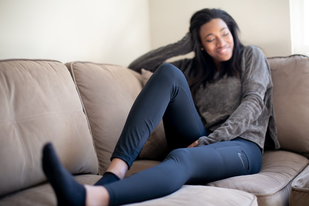 A gray long sleeves top and black leggings are worn by a woman seated on a brown couch