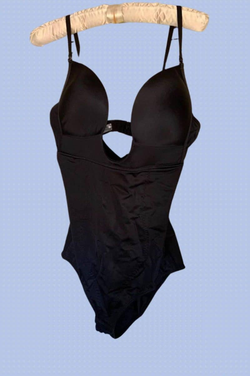 Black full-body shapewear hangs on a cream hanger placed on a purple background