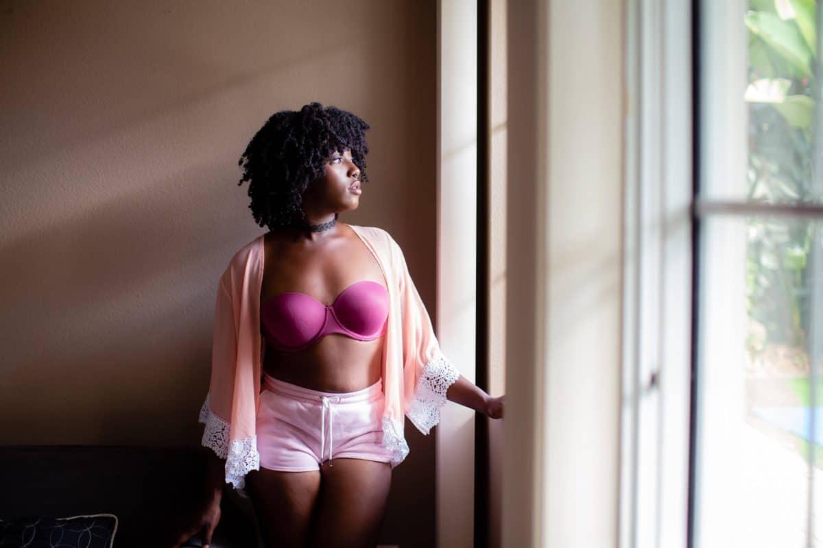 A woman in a pink strapless underwired bra and a peach cardigan wearing pink shorts staring outside the window