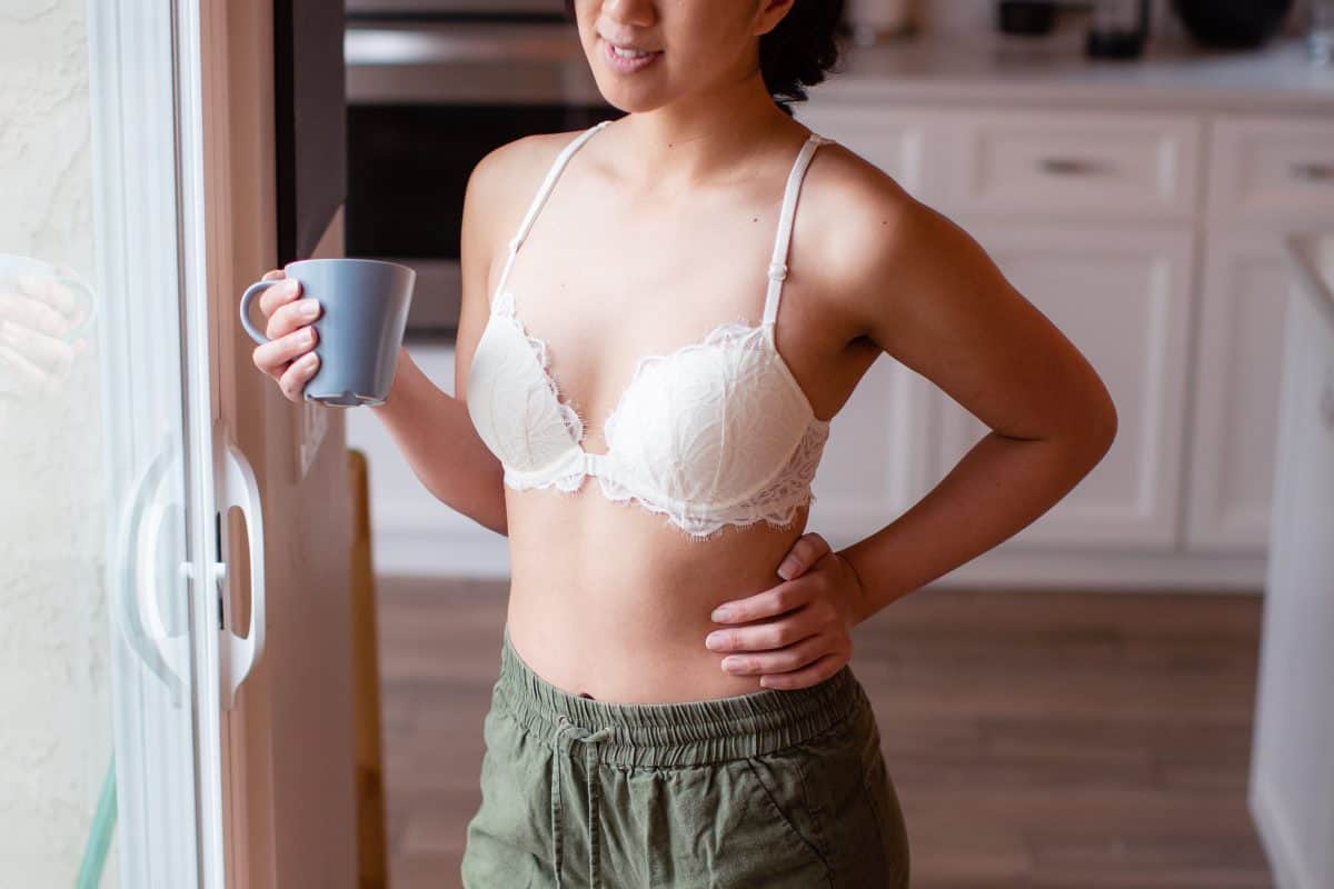 A woman in a white underwired bra and green shorts holds a blue mug while standing near a window