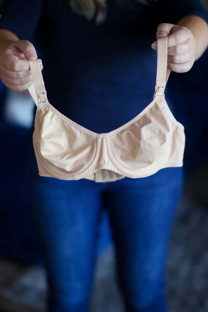 A person in a blue t-shirt and denim jeans holds a brown nursing bra with underwire and front clips