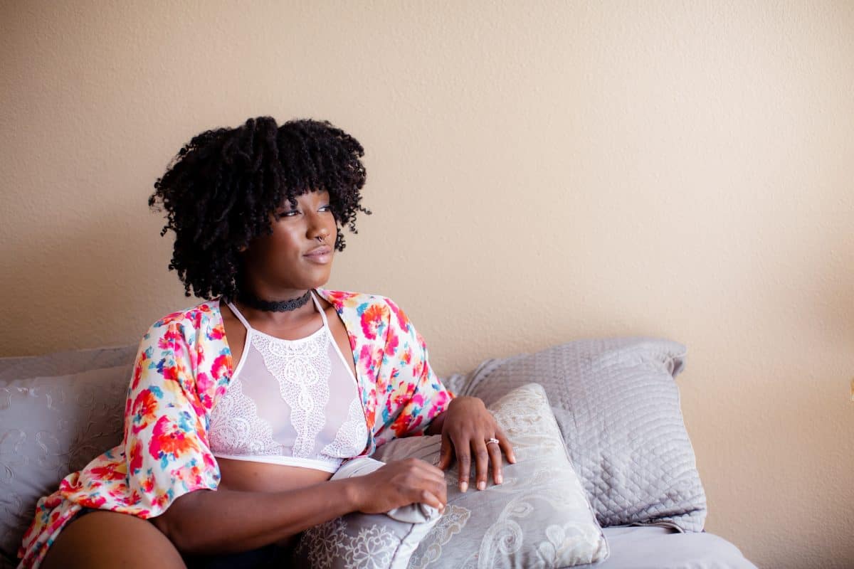 A white high-neck bralette and colorful shawl are worn by a woman seated on a gray couch