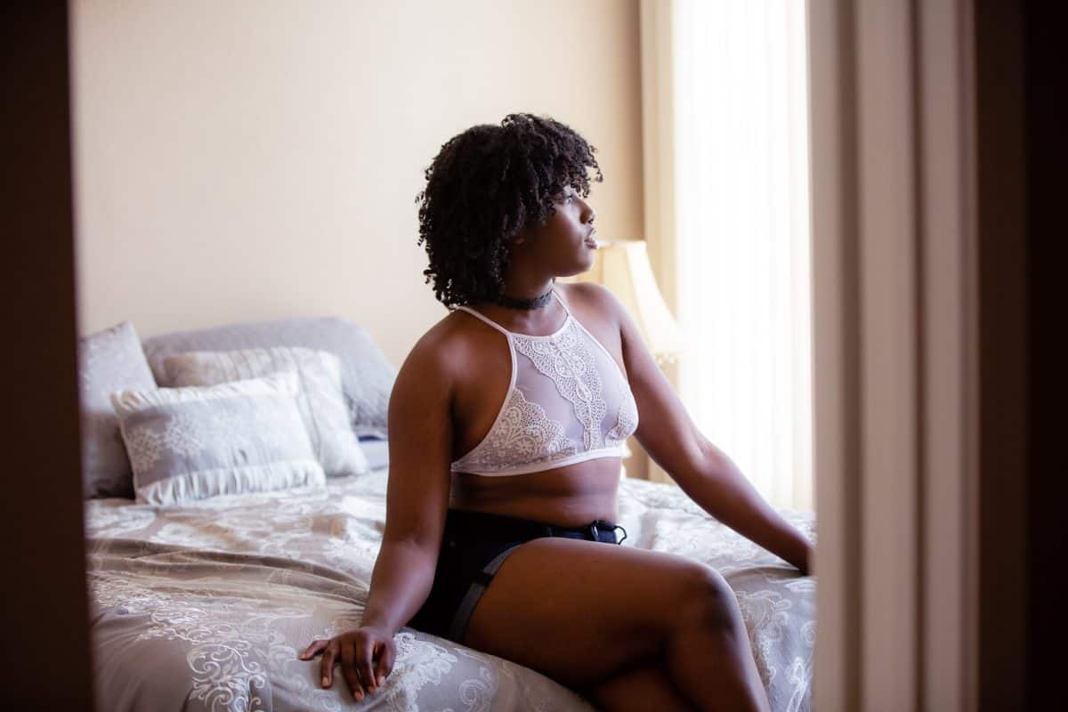 A woman wearing a white high-neck bralette and denim shorts is seated in the bed