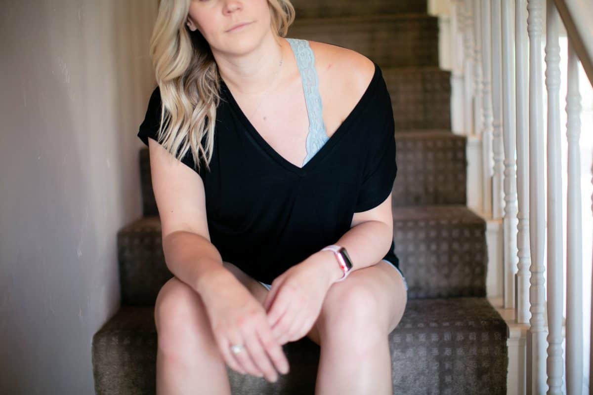 A black v-neck blouse on top of a blue lace bralette and denim shorts are worn by a woman seated on the stairs