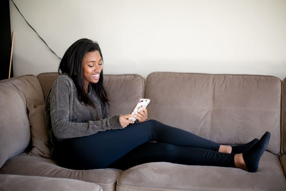 A woman wearing gray long sleeves and black leggings using a smartphone while seated on a brown couch