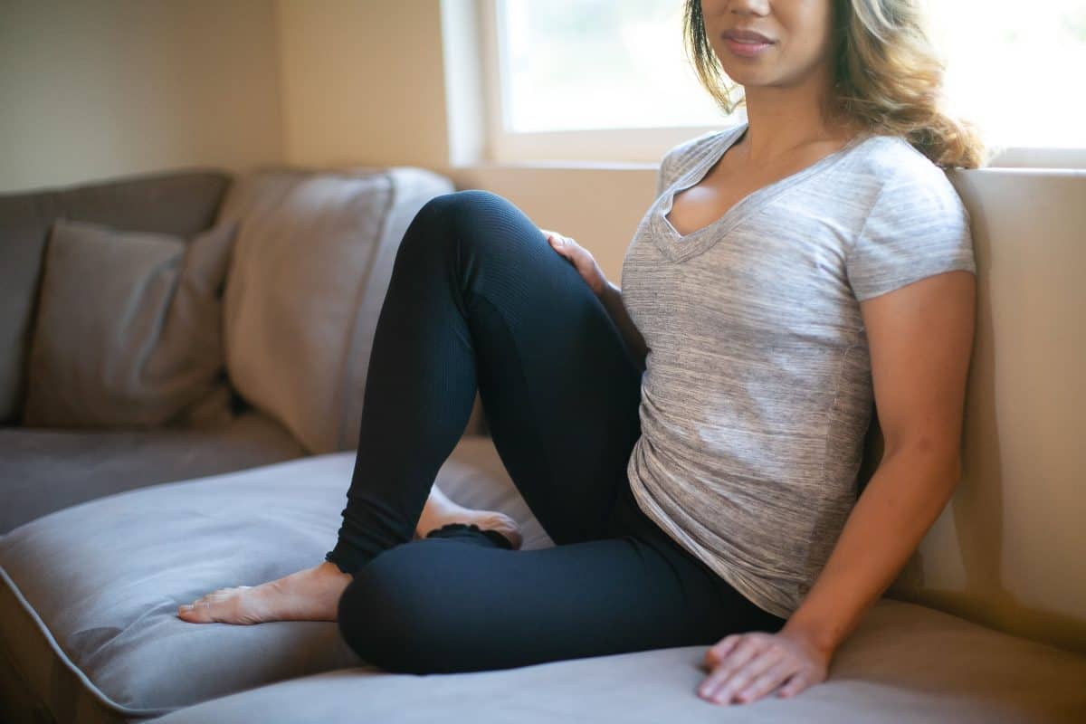 A woman wearing a gray v-neck shirt and black leggings is seated on a brown couch
