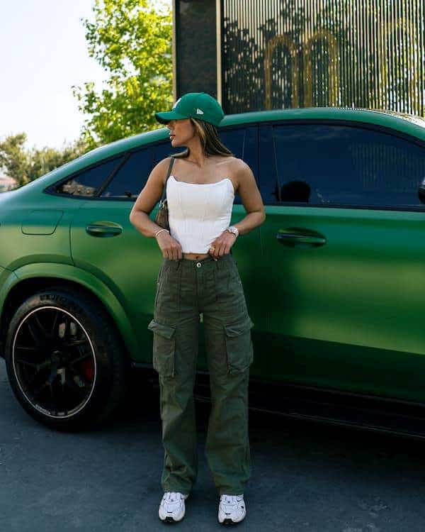 Woman wearing green cargo pants, a green cap and a white top while posing beside a green car outdoors