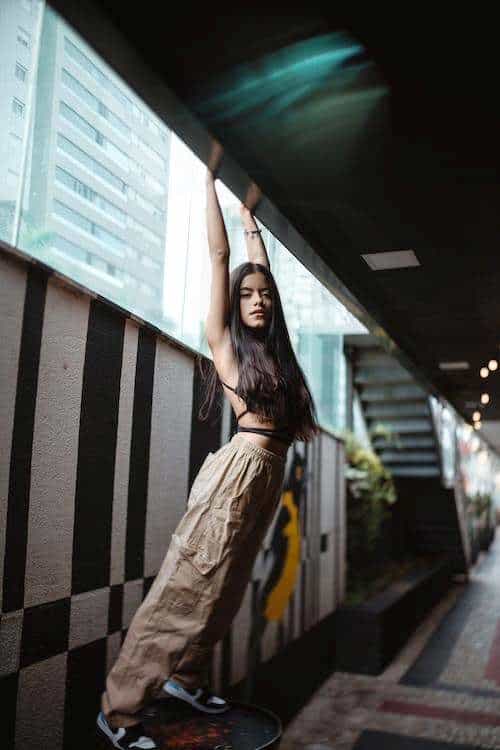 Woman wearing brown cargo pants and a strappy crop top while raising her arms high towards the roof of a parking lot for a shoot