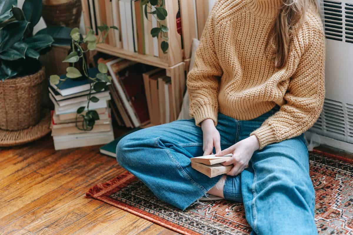 A woman sitting on a floor holding a small book wearing a brown knitted sweater and jeans