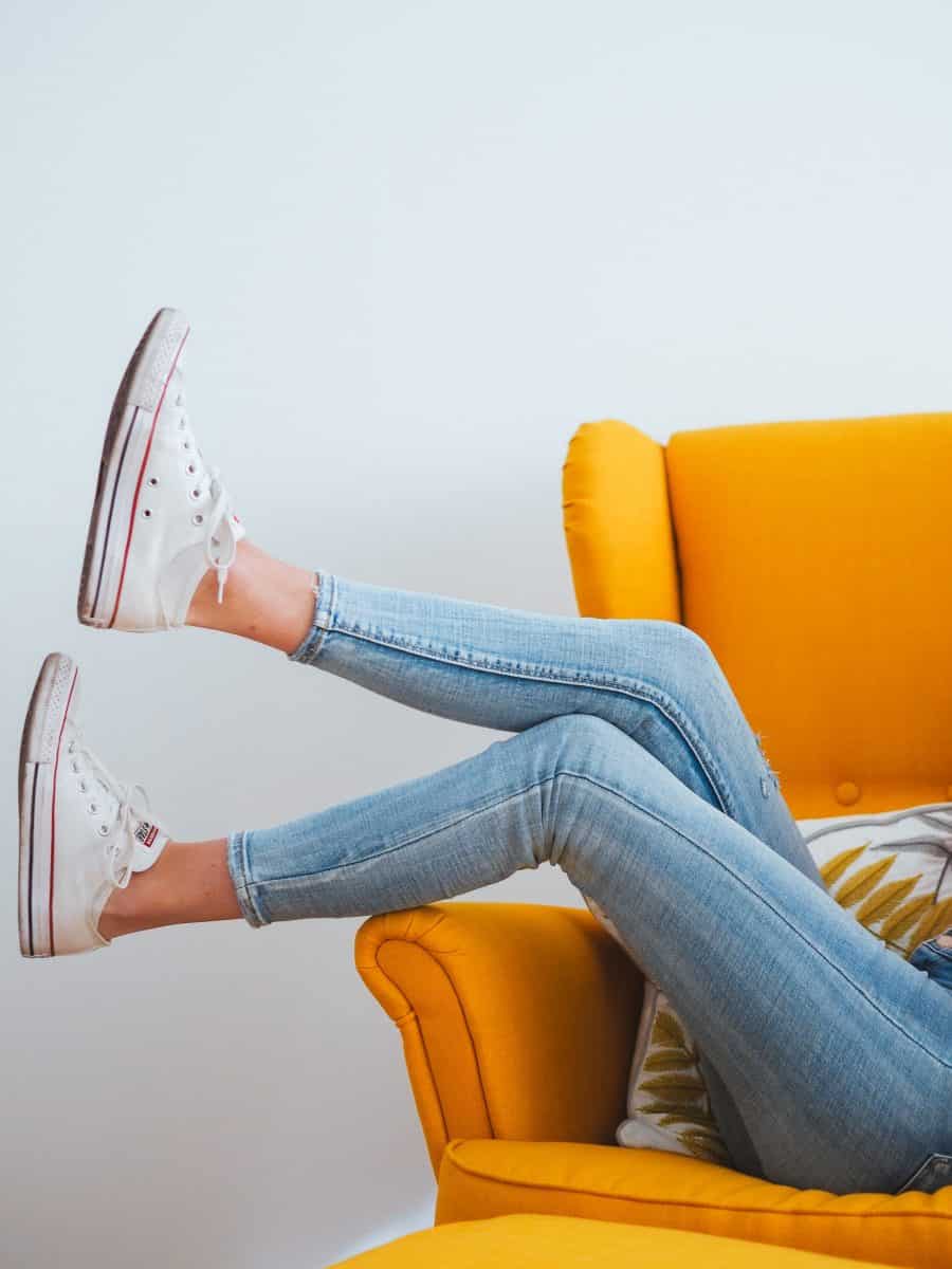 A woman's legs shown wearing white sneakers and tight blue jeans while sitting on a yellow sofa