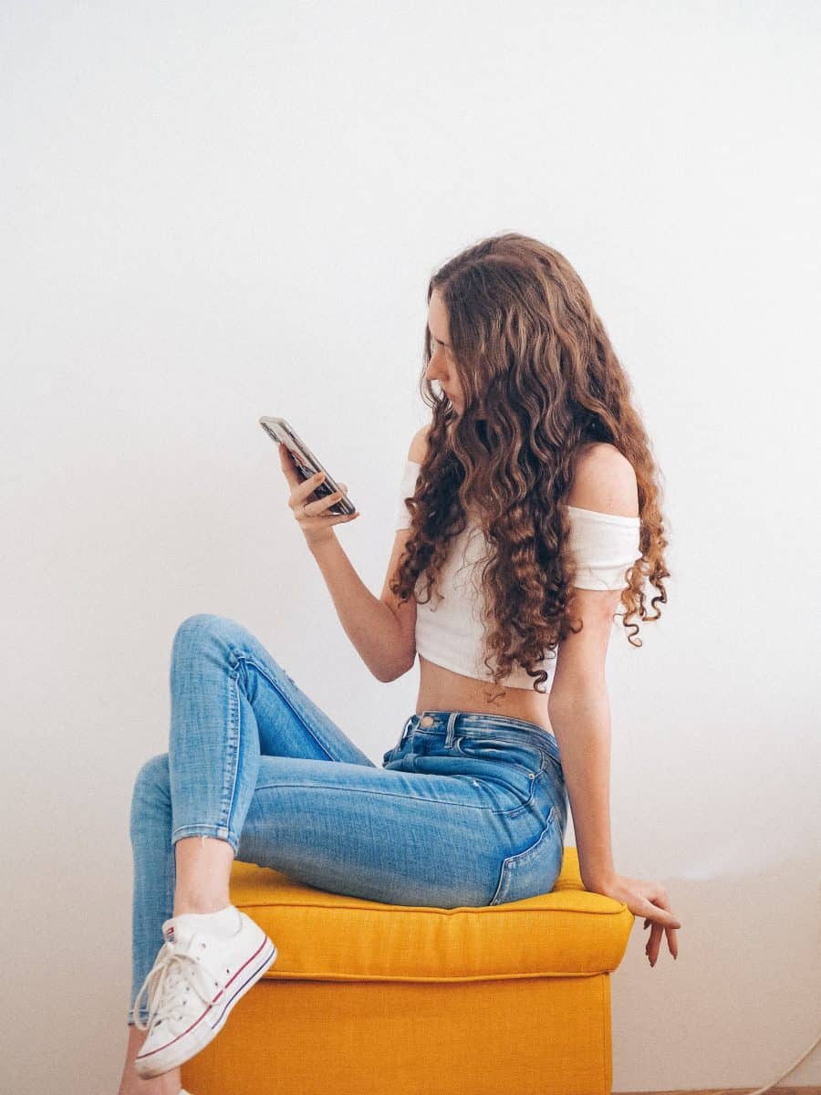 A curly haired woman holding a phone sitting on a yellow chair wearing tight jeans and crop top off shoulder dress